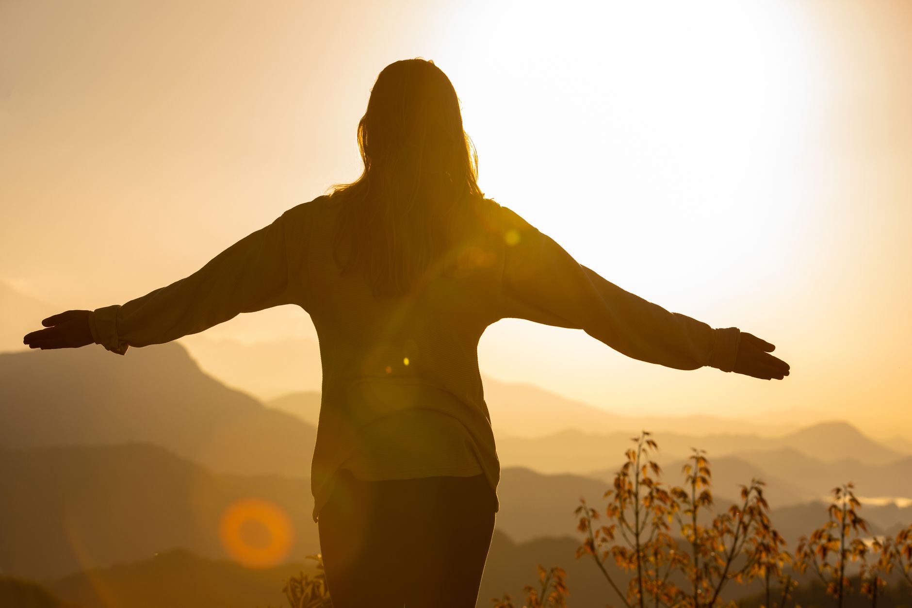 A woman stands with arms open, embracing the sunrise in a mountainous landscape, creating a tranquil silhouette.